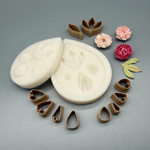 Pode incluir: Dois moldes de silicone em tom esbranquiçado com impressões de pétalas, acompanhados por um conjunto de cortadores de pétalas de plástico castanho de várias formas e tamanhos, além de três pequenos modelos de flores acabados em rosa e vermelho.