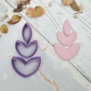 May include: A close-up shot features two sets of earring components. One set is a light purple polymer clay cutter, and the other is a pink clay earring. The components are in a layered, floral design. Dried flowers and petals are scattered around the earrings.