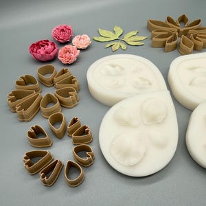 May include: A collection of tan plastic petal cutters, white silicone molds for flower shapes, and several small pink and light pink artificial peony flowers arranged on a flat surface. The set includes various sizes of teardrop and leaf-shaped cutters.