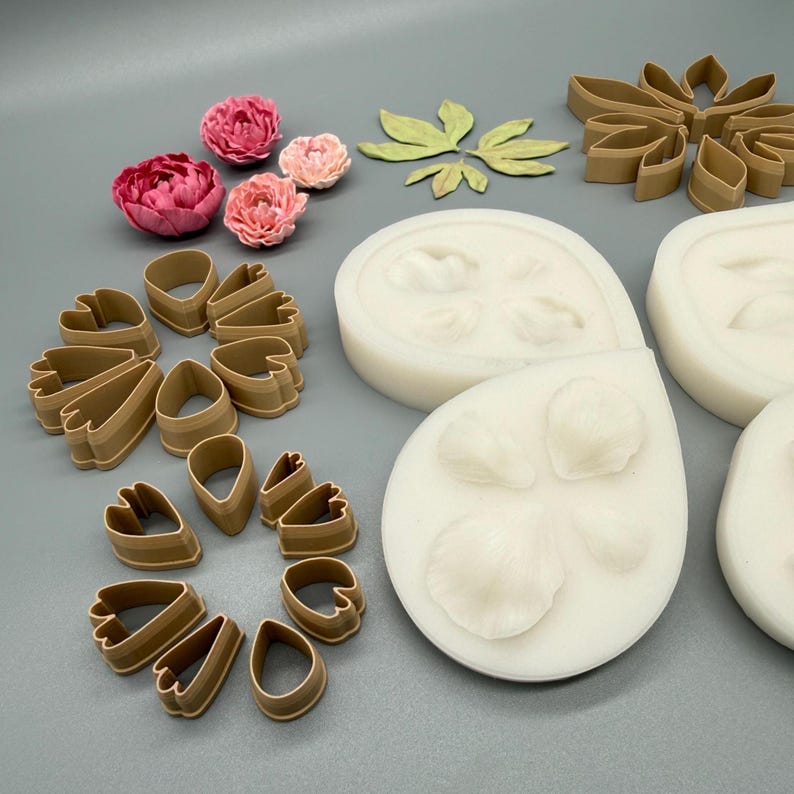 May include: A collection of tan plastic petal cutters, white silicone molds for flower shapes, and several small pink and light pink artificial peony flowers arranged on a flat surface. The set includes various sizes of teardrop and leaf-shaped cutters.