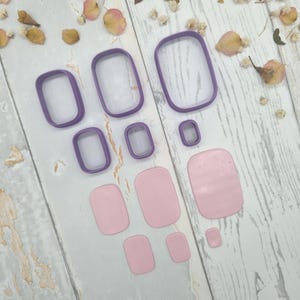 May include: A set of purple rectangular cookie cutters in various sizes arranged alongside matching light pink rectangular clay shapes. The cutters feature rounded corners and open centers, while the clay pieces are solid, smooth, and flat.