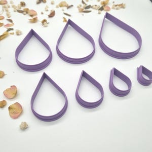 May include: A set of six lavender-colored teardrop-shaped cookie cutters arranged in two rows. The cutters vary in size, featuring a smooth, rounded base and a pointed top, constructed from a rigid plastic material with a defined cutting edge.