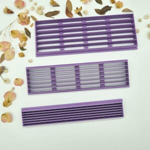 Puede incluir: Tres herramientas de repostería rectangulares moradas con listones horizontales, posiblemente para decorar pasteles, se muestran sobre una superficie blanca. Flores y hojas secas están esparcidas alrededor de las herramientas.