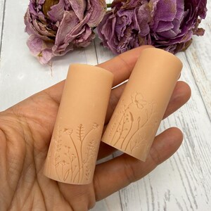 以下が含まれることがあります： ピーチ色の円筒形ワックスピラー2本。表面には野草や茎のボタニカルな線画がエンボス加工されています。滑らかでマットな質感のワックス素材は淡いオレンジ色で、表面全体に繊細で複雑な花の彫刻が施されています。