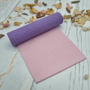 Könnte beinhalten: Eine texturierte violette zylindrische Walze, die auf einem flachen, rechteckigen Blatt aus hellrosa Material liegt. Beide Gegenstände weisen eine konsistente, feinkörnige, poröse Oberflächenstruktur auf, die einen Kontrast zu den glatten Formen bildet.