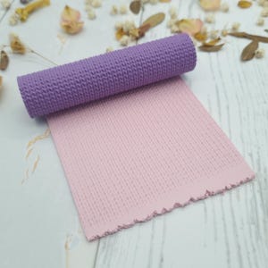 May include: A rolled-up purple textured mat rests on a flat pink mat with a matching texture. The mats have a knitted pattern. The background is a light wood surface with dried flowers.