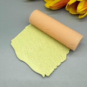 May include: A light peach-colored rolling pin rests on a textured, pale green clay surface. The clay has a rough, uneven texture. The background is a solid gray color with a few yellow and orange flower petals.