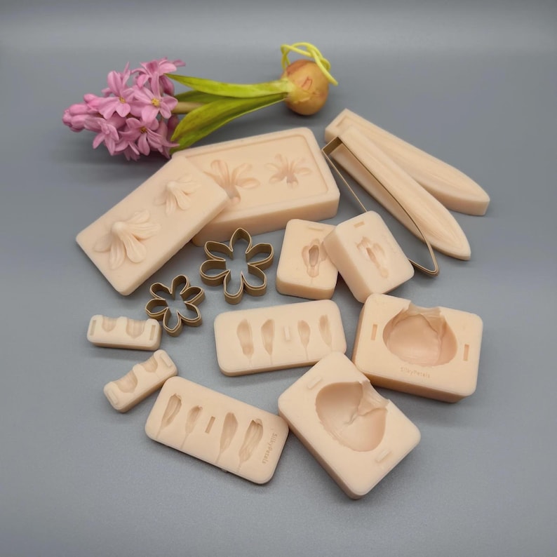 K&ouml;nnte beinhalten: Eine Sammlung beiger Silikonformen und bl&uuml;tenf&ouml;rmiger Ausstecher. Die Formen weisen Blumen- und Federdesigns auf. Eine rosa Hyazinthenbl&uuml;te und eine Zwiebel befinden sich im Hintergrund. Die Artikel sind auf einer grauen Oberfl&auml;che angeordnet.