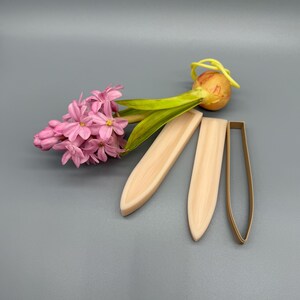 K&ouml;nnte beinhalten: Arrangement mit rosa Hyazinthenbl&uuml;ten, einer kleinen Zwiebel mit gr&uuml;nen Bl&auml;ttern, zwei Holzformen in Blattform und einem metallenen Ausstecher in Blattform. Der Hintergrund ist einfarbig grau.