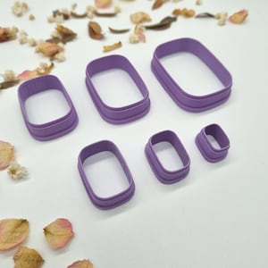 Puede incluir: Un conjunto de seis cortadores de galletas rectangulares morados de varios tamaños. Los cortadores son de plástico y tienen un borde liso y redondeado. Están dispuestos sobre una superficie blanca, con flores secas y pétalos esparcidos.