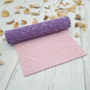 Può includere: Un mattarello cilindrico viola con un motivo floreale e a viticci in rilievo, posizionato sopra un foglio rettangolare piatto di materiale rosa chiaro che presenta lo stesso disegno di fiori e viticci impresso sulla superficie.