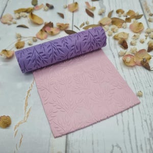 Könnte beinhalten: Eine zylindrische violette Teigrolle mit eingraviertem Blattmuster und eine quadratische Platte aus hellrosa Ton, die dasselbe geprägte Blattdesign aufweist. Die Teigrolle liegt horizontal am oberen Rand der flachen Tonplatte.