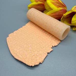 Peut inclure: Un rouleau à texture cylindrique en silicone translucide avec un motif en grille, reposant sur un morceau d'argile plat de couleur pêche clair embossé avec le même motif de grille carrée, placé près de pétales de tulipes jaunes et orange.