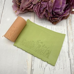 Op de afbeelding: Een cilindervormige, terracotta-kleurige kleiroller met een gegraveerd botanisch patroon van wilde bloemen en stengels, geplaatst naast een rechthoekige plak lichtgroene klei die dezelfde bloemenafdruk in het oppervlak heeft.