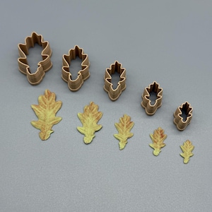 May include: A set of five light brown plastic oak leaf cookie cutters arranged in a row, accompanied by five corresponding oak leaf shapes in graduated sizes, featuring a yellow and green gradient color pattern on their textured surfaces.