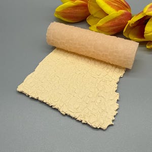 May include: A textured, beige material, rolled and unrolled, with a pattern of small, irregular shapes. The material is set against a gray background, with yellow and orange tulips in the upper right corner.