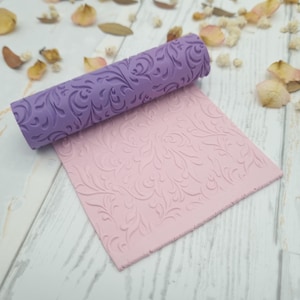 Pode incluir: Um rolo roxo com um padrão floral repousa sobre uma folha rosa texturizada. O rolo é cilíndrico e apresenta um design em relevo repetido. A folha rosa também tem o mesmo padrão floral.
