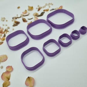 May include: A collection of seven purple square and rectangular cookie cutters in different sizes. The cutters are made of plastic and have a rounded edge. The image is taken from a slightly elevated angle, with a white background and scattered dried flower petals.