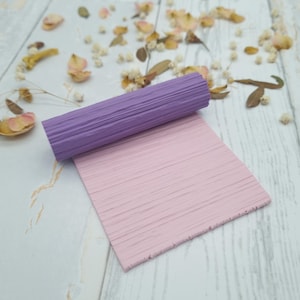 Könnte beinhalten: Ein zylindrisches, violettes Nudelholz mit einer texturierten Oberfläche aus horizontalen Linien, kombiniert mit einer flachen, quadratischen, hellrosa Platte, die ebenfalls eine durchgehende horizontale Linienstruktur aufweist.