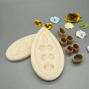 Op de afbeelding: Een verzameling bakbenodigdheden voor het maken van bloemdecoraties. De afbeelding toont een beige siliconen mal in druppelvorm, bloemvormige uitstekers en verschillende handgemaakte viooltjes in geel, blauw en wit.