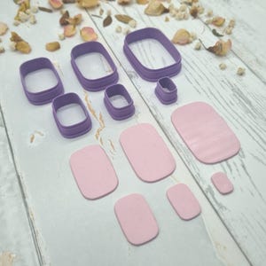 May include: A set of purple rectangular cookie cutters in various sizes alongside matching light pink rectangular clay shapes. The cutters feature rounded corners and a sturdy rim, while the corresponding clay pieces are smooth and flat.