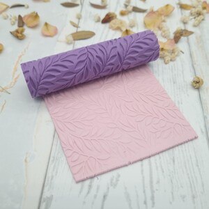 Op de afbeelding: Een paarse cilindrische deegroller met een verhoogd bladpatroon en een bijpassend lichtroze vierkant vel materiaal met hetzelfde reliëf bladontwerp, dat de textuur toont die door de roller op het platte oppervlak is gecreëerd.