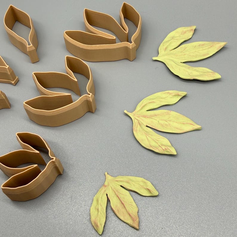 May include: A collection of tan plastic leaf-shaped cookie cutters and three corresponding light green leaf-shaped cutouts with visible veining details. The cutters feature open centers and raised edges for shaping dough or clay into botanical forms.