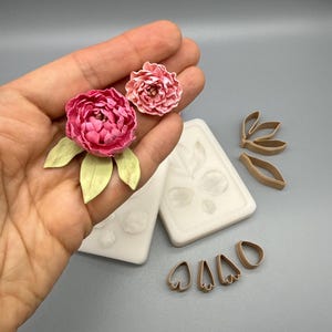 Puede incluir: Dos peonías rosas con hojas verde claro, dos moldes de silicona blancos con impresiones florales y varios cortadores de pétalos de plástico marrón. Las flores presentan pétalos en capas y se muestran junto a las herramientas de creación.
