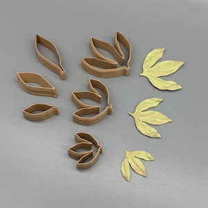 May include: A collection of light brown plastic cookie cutters in various leaf shapes, including single-lobed and multi-lobed designs, alongside three corresponding pale yellow leaf-shaped cutouts made from a flat, pliable material.