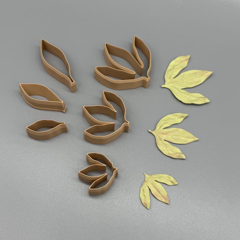 May include: A collection of light brown plastic cookie cutters in various leaf shapes, including single-lobed and multi-lobed designs, alongside three corresponding pale yellow leaf-shaped cutouts made from a flat, pliable material.
