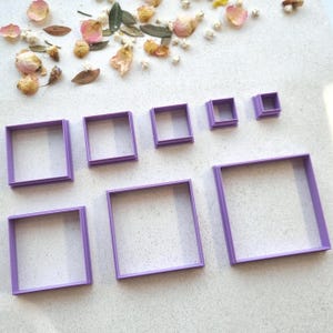 May include: A collection of seven square cookie cutters in different sizes. The cutters are a solid lavender colour and are arranged from largest to smallest. Dried flowers and leaves are scattered around the cutters on a white surface.