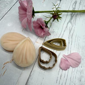 Può includere: Una natura morta con due fiori rosa chiaro, due saponi a forma di petalo, due taglia biscotti a forma di petalo e due petali rosa. Gli oggetti sono disposti su una superficie di legno chiaro.