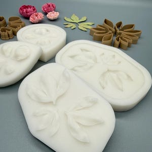 May include: White silicone molds featuring embossed leaf and petal patterns, accompanied by brown plastic cutters in matching shapes and several small, realistic pink and red artificial flower blossoms arranged on a flat surface.