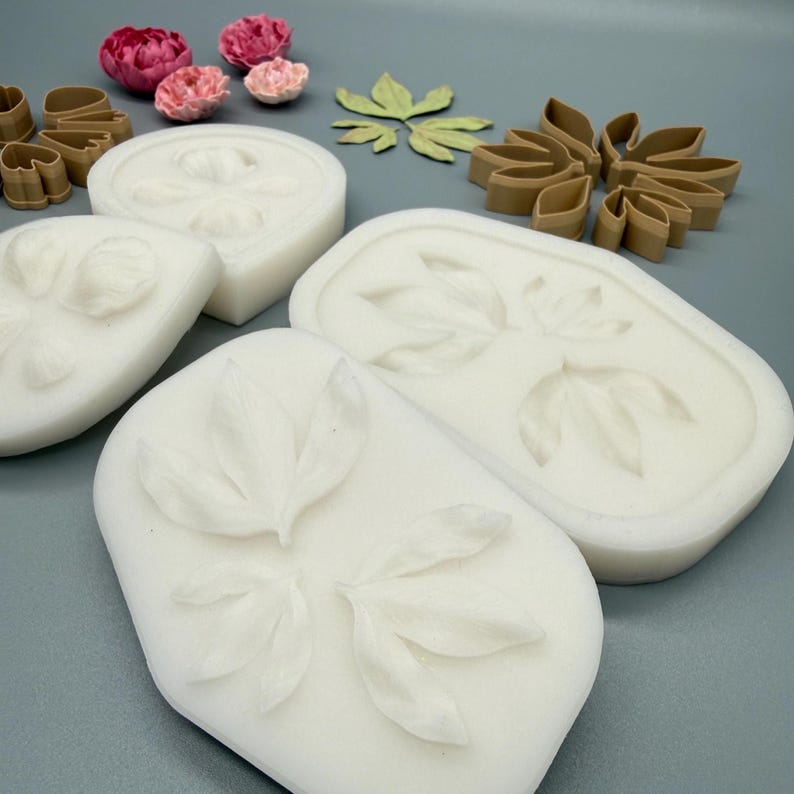 May include: White silicone molds featuring embossed leaf and petal patterns, accompanied by brown plastic cutters in matching shapes and several small, realistic pink and red artificial flower blossoms arranged on a flat surface.