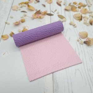 Könnte beinhalten: Ein quadratisches Blatt aus hellrosa strukturiertem Material mit einem darauf liegenden, gerollten Abschnitt aus violettem, strukturiertem Material. Beide Teile weisen ein durchgehendes, geprägtes Narbenmuster auf ihren Oberflächen auf.