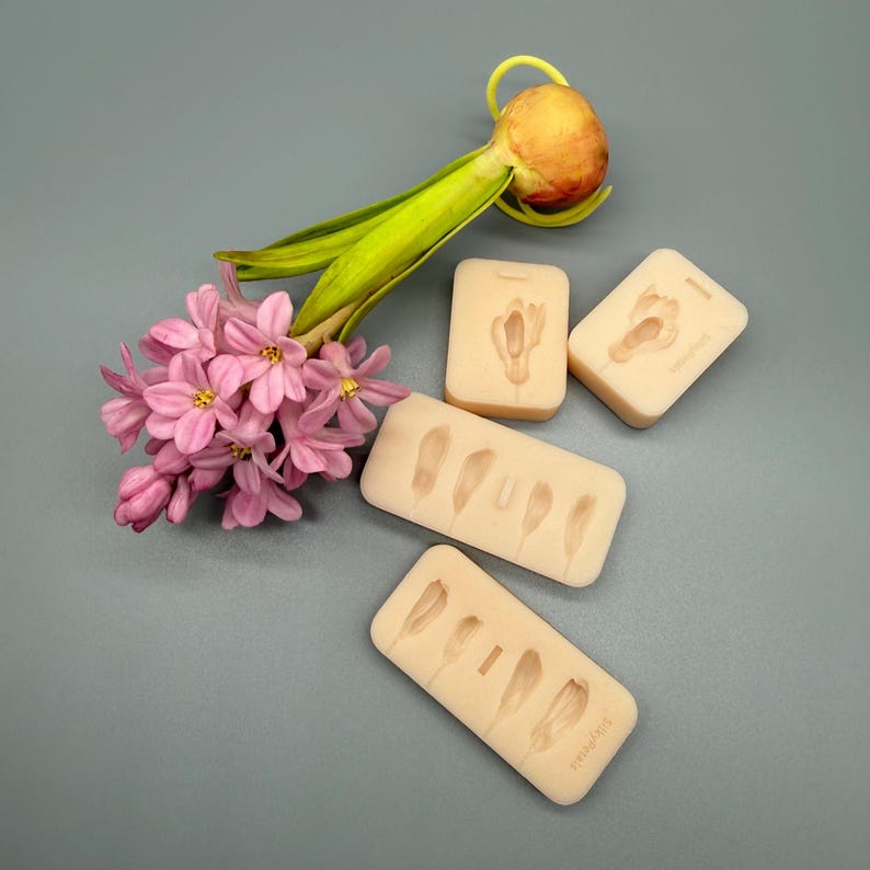 K&ouml;nnte beinhalten: Ein Stillleben mit rosa Hyazinthenbl&uuml;ten, einer Zwiebel und mehreren beigefarbenen Silikonformen mit Blumen- und Federdesigns. Die Formen sind rechteckig und haben detaillierte Abdr&uuml;cke. Der Hintergrund ist einfarbig grau.