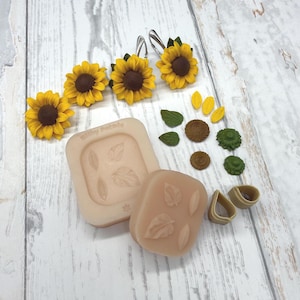 Könnte beinhalten: Ein Set mit Bastelbedarf zum Thema Sonnenblume, bestehend aus drei fertigen Sonnenblumen-Ohrringen, zwei beigen Silikonformen mit Blattabdrücken, zwei beigen Blütenblatt-Ausstechern sowie verschiedenen losen Miniatur-Sonnenblumen-Elementen wie Blütenblättern, Kernen und grünen Blättern.