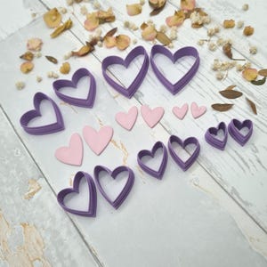 Op de afbeelding: Een assortiment paarse hartvormige koekjessnijders en roze hartvormige fondantdecoraties zijn gerangschikt op een licht oppervlak. Gedroogde bloemen en bladeren zijn rond de harten verspreid, waardoor een decoratieve presentatie ontstaat voor bakken en knutselen.