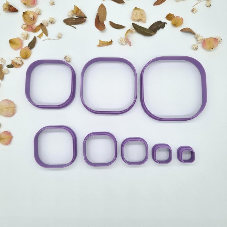 May include: A collection of seven purple square cookie cutters in different sizes. The cutters are arranged in two rows, with the largest at the top and the smallest at the bottom. The background is white with dried flowers and leaves scattered around.