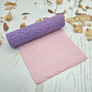 Op de afbeelding: Een getextureerde, lavendelkleurige deegroller ligt op een roze, getextureerd vel. De deegroller en het vel hebben een vergelijkbaar patroon, waardoor een samenhangend ontwerp ontstaat. De items worden waarschijnlijk gebruikt voor knutselen of bakken. Gedroogde bloemblaadjes zijn verspreid.