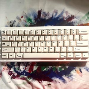 May include: A white mechanical keyboard with off-white keycaps. The keyboard has a standard QWERTY layout with Japanese characters on some keys. The background features colorful paint splatters.