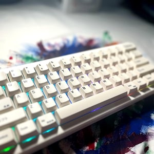 May include: A white mechanical keyboard with illuminated keys. The keyboard has a compact layout with white keycaps and a white frame. The keys are backlit with a colorful glow. The keyboard is on a white surface.
