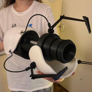 May include: A white and black prop device with a large black barrel and articulated arms. The device has a curved white base. The prop is held by a person wearing a white t-shirt. Likely from a science fiction or video game.