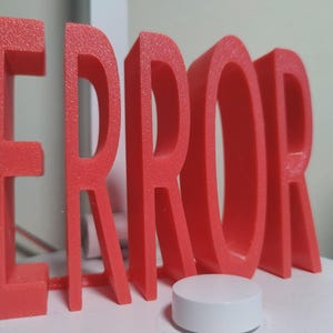 以下が含まれることがあります： 3Dプリントされたコーラル色の「ERROR」という単語が白い表面に立っています。小さな白い円筒形の物体が単語の前にあります。文字は太くブロック状で、表面はざらざらしています。