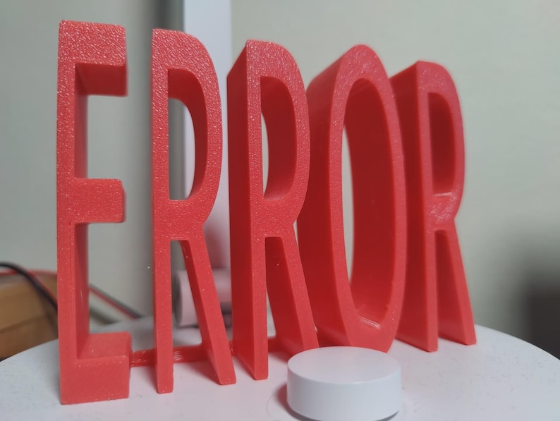 以下が含まれることがあります： 3Dプリントされたコーラル色の「ERROR」という単語が白い表面に立っています。小さな白い円筒形の物体が単語の前にあります。文字は太くブロック状で、表面はざらざらしています。