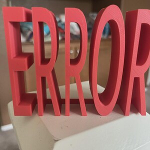 以下が含まれることがあります： 「ERROR」という文字が書かれた赤い3Dプリントのサイン。文字は太く、白い表面に立っています。このサインは、大胆で人目を引く装飾品です。