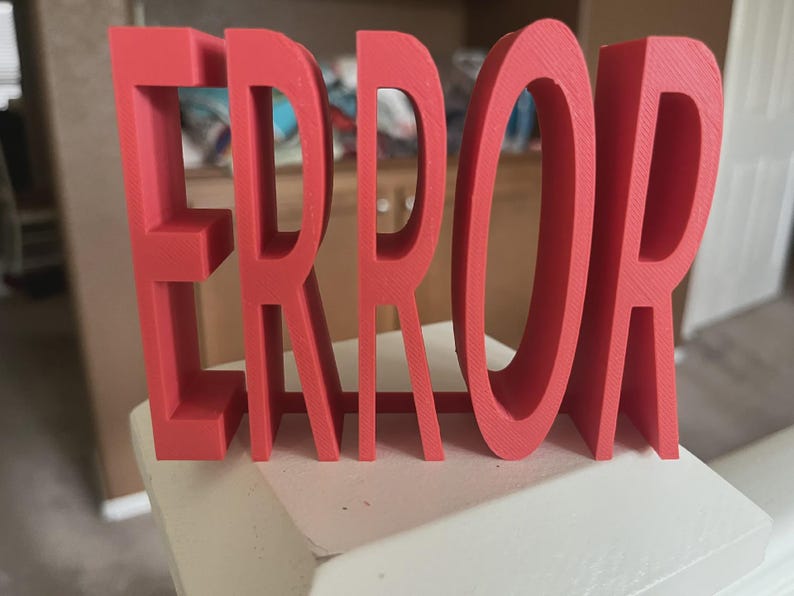 以下が含まれることがあります： 「ERROR」という文字が書かれた赤い3Dプリントのサイン。文字は太く、白い表面に立っています。このサインは、大胆で人目を引く装飾品です。