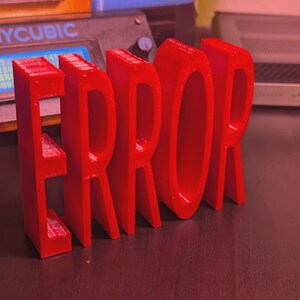 以下が含まれることがあります： 鮮やかな赤色の3Dプリントされた「ERROR」の文字が、暗い表面に立っています。文字は太くブロック状で、光沢のある仕上がりです。背景には3Dプリンターとノートパソコンが見えます。