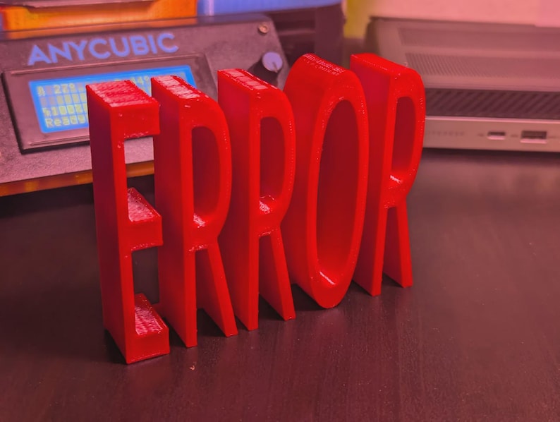 以下が含まれることがあります： 鮮やかな赤色の3Dプリントされた「ERROR」の文字が、暗い表面に立っています。文字は太くブロック状で、光沢のある仕上がりです。背景には3Dプリンターとノートパソコンが見えます。
