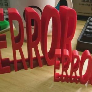 Puede incluir: Dos palabras "ERROR" rojas impresas en 3D, de diferentes tamaños. La más grande está de pie, la más pequeña delante. Las letras son gruesas y el acabado es mate. Un teclado y una taza blanca están en el fondo.